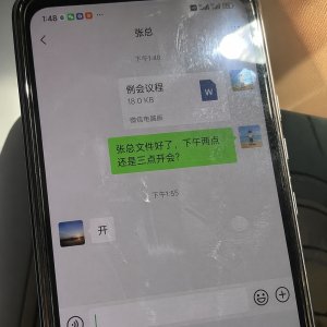 一张真实拍摄的手机照片，手机屏幕显示微信聊天界面（中文对话），有聊天气泡、文件（Word）附件，界面为绿色和灰色气泡，屏幕上有明显指纹、油渍、划痕，玻璃有强烈反光，光源直射形成眩光，画面略微倾斜，手持拍摄，环境自然光，不完美构图，真实感强，生活化随手拍，高细节。 对话内容为老板和员工的对话，员工发文件（文件名&rdquo;例会议程&ldquo;）给张总，并说：张总文件好了，下午两点还是三点开会？张总说：开。